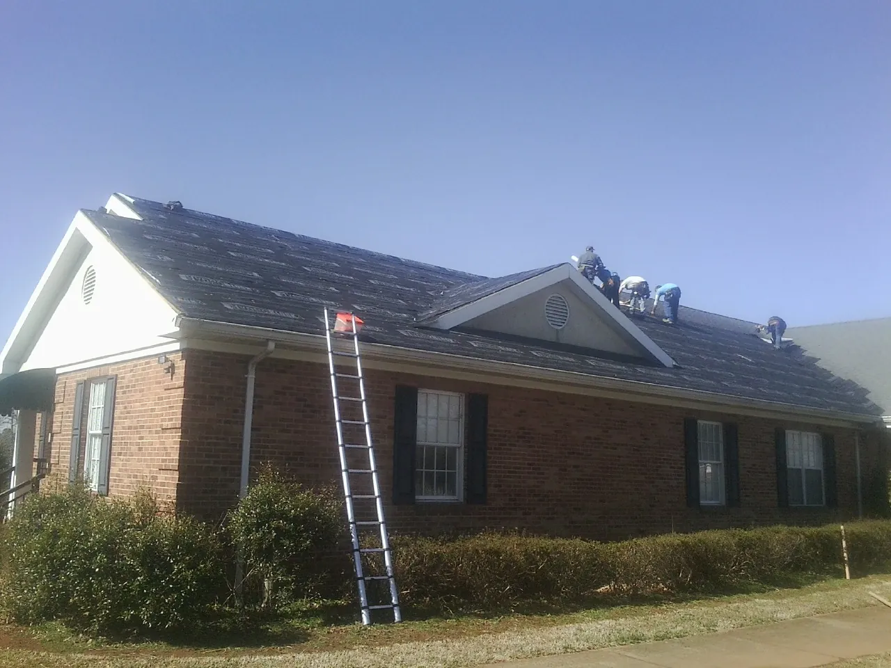 Crew tearing shingles off a brick ranch roof with a ladder set — Emergency Roof Repair work by our crew in Sodus