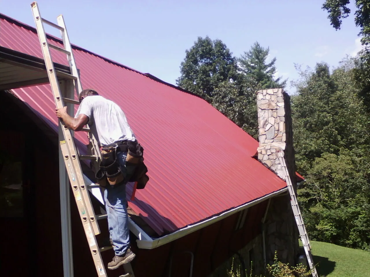 Roofer on a ladder beside a red metal roof and stone chimney — Soffit & Fascia Repair by Roofing Brothers in Sodus